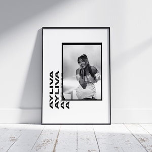 May include: Monochrome framed print of a woman singing into a microphone. The print has the word "AYLIVA" stacked vertically on the left. The image is displayed on a white wall with a wooden floor.