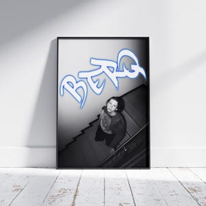 May include: Black and white framed print featuring a person looking up from a staircase. The word "BERG" is written in blue graffiti-style lettering above the person. The image is displayed on a white wall.