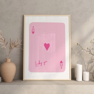Może przedstawiać: Oprawiony różowy nadruk przedstawiający kartę do gry, Asa Kier. Karta ma duże serce na środku i mniejsze serca w rogach. Słowa "Lucky you" są napisane kursywą.