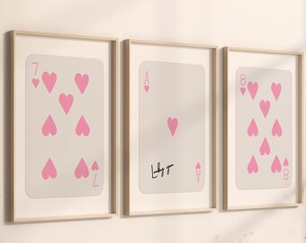 May include: Three framed playing card prints. The cards are light beige with pink hearts. The cards are the 7 of hearts, the Ace of hearts with a signature, and the 8 of hearts. The frames are light wood.