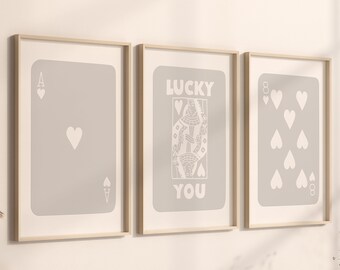 May include: Three framed art prints featuring playing card designs. The prints include an Ace of Hearts, a Queen of Hearts with the text "LUCKY YOU", and an Eight of Hearts. The cards are light gray with white details.