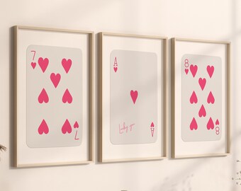 May include: Three framed playing card prints. The cards are the 7 of hearts, Ace of hearts, and 8 of hearts. Each card has a light background with pink heart symbols. The frames are light wood.