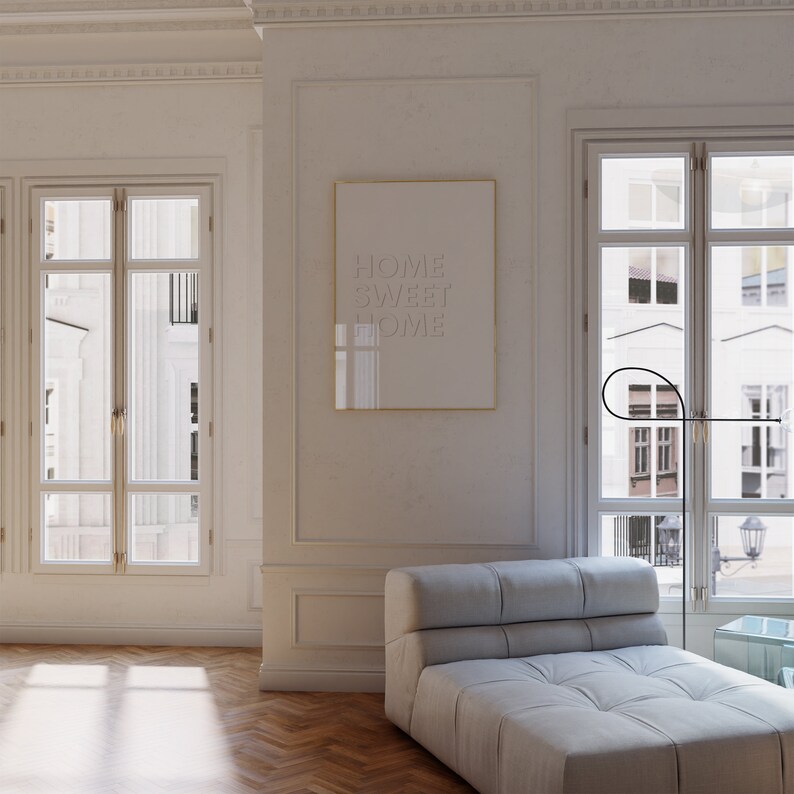 May include: A white framed poster with the text "HOME SWEET HOME" hanging on a white wall in a living room. A white couch with a tufted design is in the foreground. The room has large windows with white trim and a wooden floor.