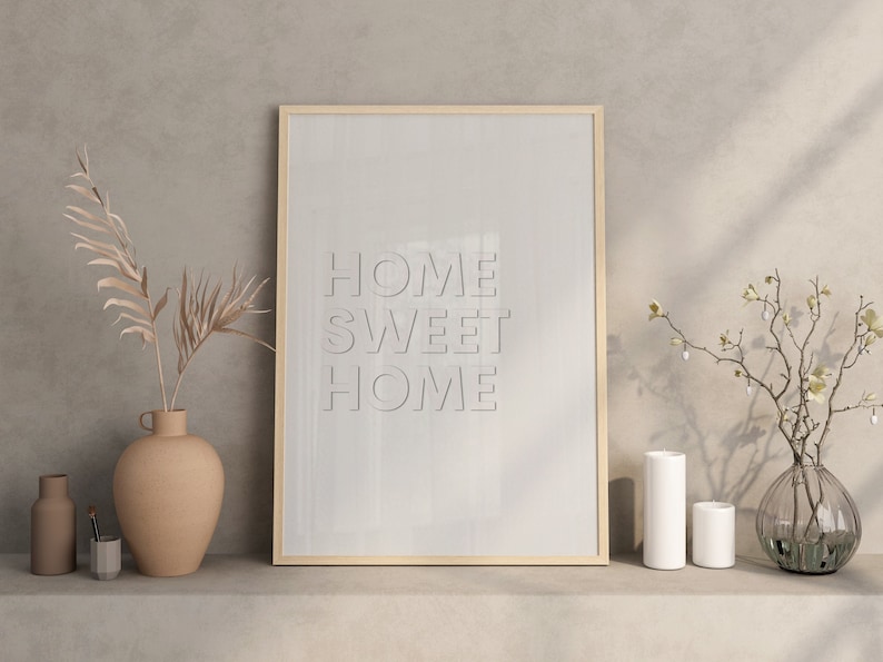 May include: A framed print with the text "HOME SWEET HOME" in a light gray color on a white background. The print is on a shelf with a vase of dried palm leaves, two candles, and a vase of white flowers.
