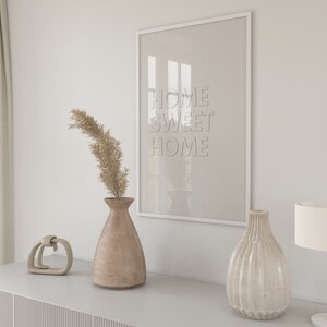 May include: A white framed picture with the text "Home Sweet Home" hanging on a white wall above a gray dresser. There are two vases, one brown and one white, and a small decorative object on the dresser. A lamp with a white shade is on the right side of the dresser.