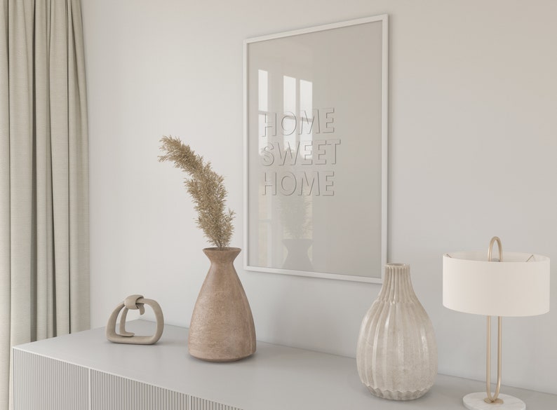 May include: A white framed picture with the text "Home Sweet Home" hanging on a white wall above a gray dresser. There are two vases, one brown and one white, and a small decorative object on the dresser. A lamp with a white shade is on the right side of the dresser.