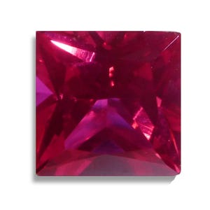 May include: A square-cut, faceted ruby gemstone. The gem is a deep, saturated red color with internal reflections. The facets create a play of light and shadow, enhancing the gem's brilliance. The gemstone is set against a white background.