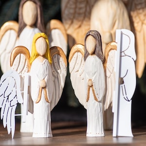 May include: Wooden angel figurines with carved details. The angels have white robes, detailed wings, and varying hair colors, including blonde and brown. Some wings have gold accents. The figurines are standing on a wooden surface.