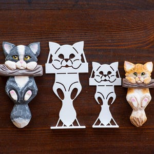 May include: Four cat-themed items on a dark wood surface. Two are carved wooden cat figures, one gray and white, the other orange and white. Two are white cat-shaped cutouts. All have a similar design, with the cats holding a log.