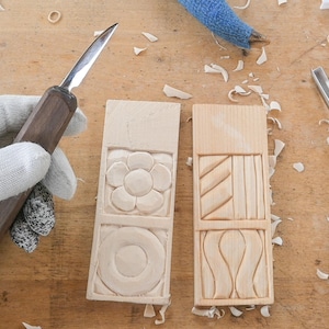 May include: Two rectangular wooden carvings with floral and geometric designs, alongside carving tools. One carving features a flower and a circle, the other has angled lines and curved shapes. The tools include a knife and a chisel.