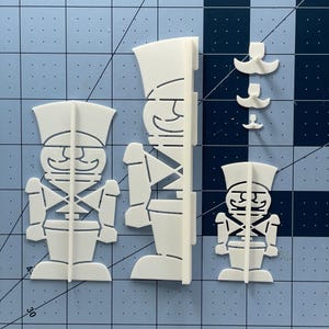 May include: Three white paper cutouts of toy soldiers. The soldiers are in different poses and have a simple design. The cutouts are on a blue and white grid background.