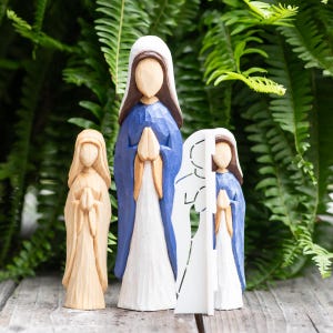 May include: Three wooden religious figurines. The tallest figure is painted blue with a white veil and a brown head. The other two figures are unpainted wood. All figures have their hands clasped in prayer. The background is green foliage.