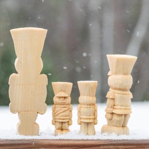 May include: Four wooden nutcracker figurines of varying sizes, standing in a row on a snowy surface. The nutcrackers are all made of unfinished wood and have a simple, traditional design.