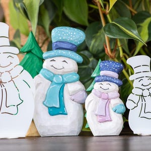 May include: Four wooden snowman figurines. Two are painted with blue hats and scarves, and two are white cutouts. A green wooden Christmas tree is between the snowmen. The snowmen have smiling faces and are suitable for holiday decor.