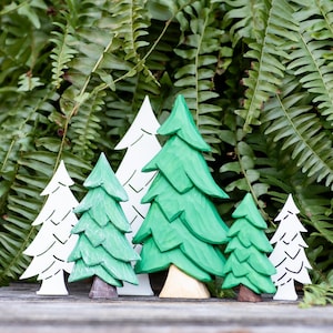 May include: A collection of wooden Christmas tree figurines in varying sizes and styles. The trees are painted in shades of green and white, with some featuring natural wood bases. The background is a lush green fern.