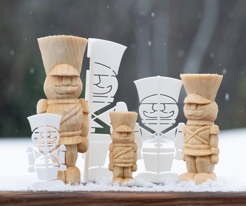 May include: Five wooden toy soldier figurines, some painted white, some unpainted, standing in a row on a snowy surface. The soldiers are all wearing hats and have a whimsical, cartoonish appearance.