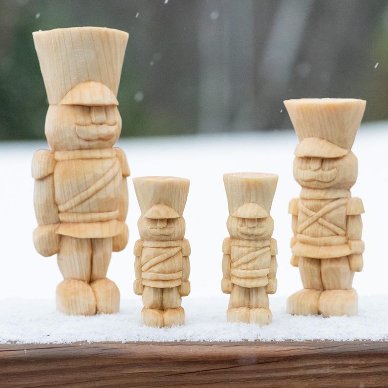 May include: Four wooden toy soldier figurines, each with a different size and a painted face. The soldiers are standing on a snowy surface.