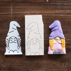 May include: A white plastic gnome template, a wooden gnome template with a pencil drawing of a gnome, and a finished painted gnome with a purple hat, yellow braids, and a pink dress.