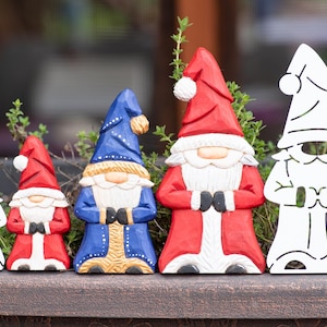 May include: A collection of five gnome figurines arranged in a row. Two are white metal silhouettes, while three are carved wooden figures painted in red, blue, and white with pointed hats, long beards, and dark gloves, standing in a line.