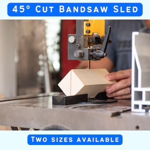 May include: A black 45 degree cut bandsaw sled holding a light-colored wooden block under a bandsaw blade. The text 45 DEGREE CUT BANDSAW SLED and TWO SIZES AVAILABLE is displayed. The sled features a V-shaped groove for positioning the wood.