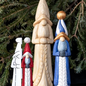May include: A collection of carved wooden figures, including a white, red, and blue gnome-like statues. The figures have elongated bodies and pointed hats. The background features green pine branches.