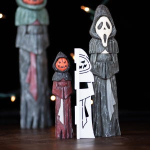 May include: Wooden Halloween figurines. The figures include a pumpkin-headed character, a Scream mask figure, and a carved wooden figure in a hooded robe. The figures are painted in shades of gray, red, and orange. The background is dark with blurred lights.