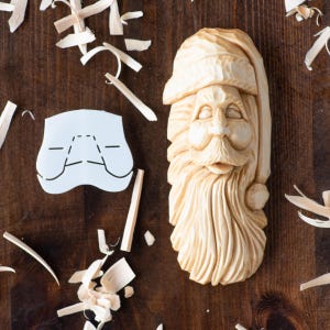 Peut inclure: Une décoration de Noël en bois sculpté représentant le Père Noël avec un chapeau blanc et une longue barbe. L'ornement est sur une surface en bois avec des copeaux de bois autour.