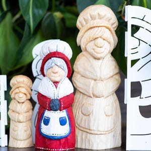 Puede incluir: Colección de figuras de madera que representan a una mujer con gorro de chef y delantal. La figura central está pintada en rojo, blanco y azul. Las otras figuras son de madera sin pintar. Un recorte decorativo blanco a la izquierda y a la derecha.