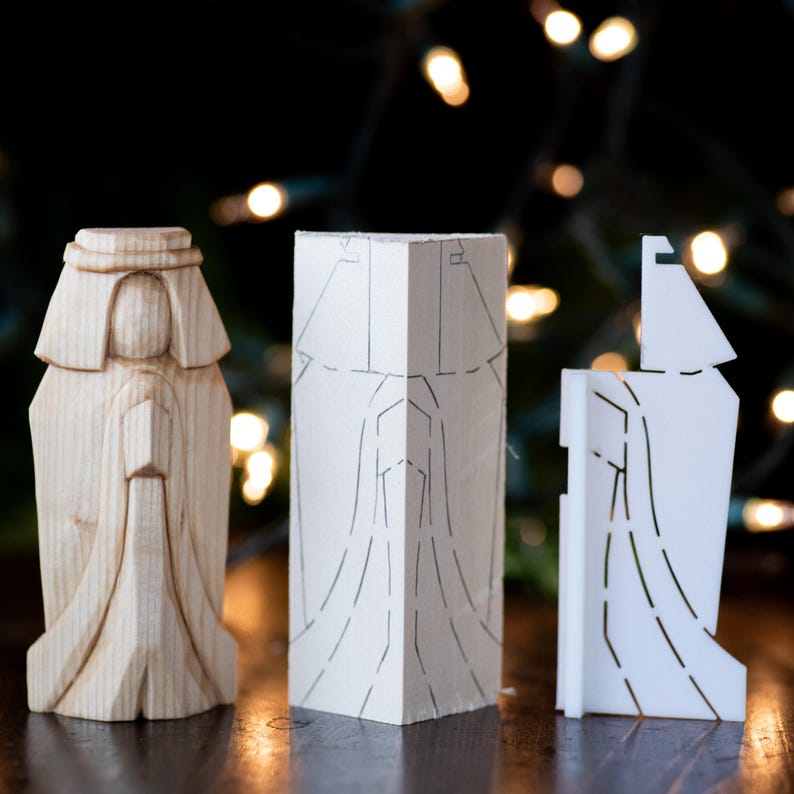 K&ouml;nnte beinhalten: Drei Holzfiguren, eine fertig und zwei unvollendet, zeigen die Phasen des Schnitzens eines Weihnachtsbaumanh&auml;ngers. Der fertige Anh&auml;nger ist eine stehende Figur mit einem langen Gewand. Die unfertigen Anh&auml;nger sind flach und zeigen die Linien der Schnitzerei.