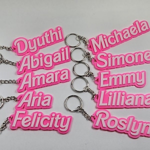 Puede incluir: Nueve llaveros rosas con letras blancas que deletrean los nombres: Dyuithi, Abigail, Amara, Aria, Felicity, Michaela, Simone, Emmy y Lilliana.