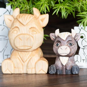 May include: Wooden cow figurines in various sizes and styles. The central cow is light brown, while the smaller one is brown with a cream-colored patch. Two white cow cutouts are also present, creating a rustic, handcrafted aesthetic.