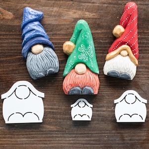 Pode incluir: Três ornamentos de gnomo de madeira com chapéus e barbas de diferentes cores. Os gnomos estão sentados em uma superfície de madeira com três recortes de casas de papel branco.