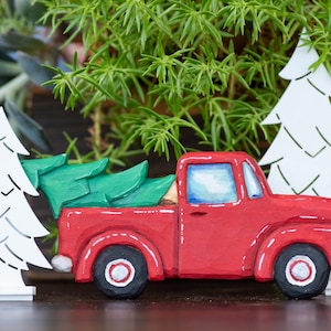 Pode incluir: Uma figura de caminhonete vermelha transportando um pinheiro verde na caçamba, ladeada por duas silhuetas de pinheiros brancas recortadas. O caminhão apresenta rodas pretas com centros brancos, janelas azuladas e acabamento pintado.