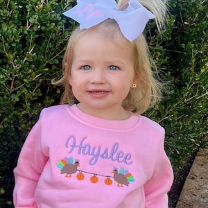 Pode incluir: Uma camisola rosa com calças de renda branca e rosa com babados na parte inferior. A camisola tem um desenho bordado de dois perus com abóboras e o nome "Hayslee".