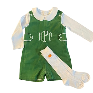 May include: A green corduroy overall with a white collared shirt and white knee-high socks with a small orange pumpkin design. The overall has a white embroidered monogram "HFP" on the front.