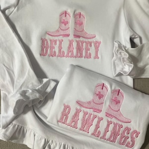 Puede incluir: Dos camisas blancas de manga larga con botas de vaquero bordadas en rosa y nombres. La camisa de arriba dice "Delaney" y la camisa de abajo dice "Rawlings".