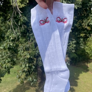 May include: A pair of white ribbed socks with a red and green embroidered bow design near the top. The socks are held up against a backdrop of green foliage and sunlight.