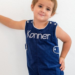 May include: A young boy wearing a blue overall with white trim and the name "Konner" embroidered on the front.