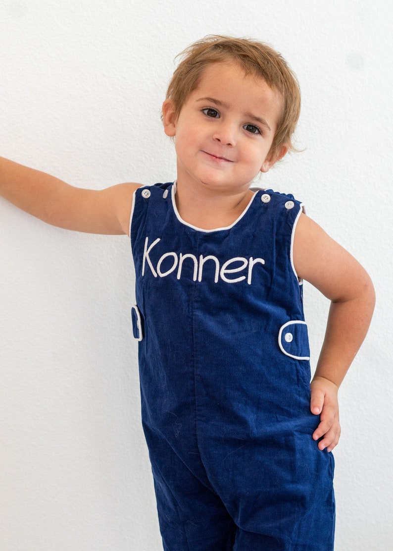May include: A young boy wearing a blue overall with white trim and the name "Konner" embroidered on the front.