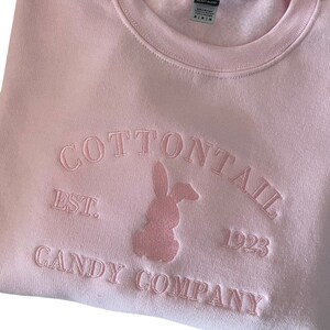 Puede incluir: Sudadera rosa con un logotipo de conejo bordado en rosa y el texto "COTTONTAIL EST. 1923 CANDY COMPANY".