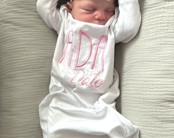 Puede incluir: Un vestido blanco de bebé con letras bordadas en rosa que dicen "ADA Dale". El vestido tiene mangas con volantes y una diadema con un lazo blanco está en la cabeza del bebé.