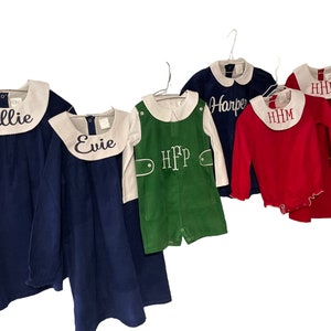 May include: Five children's clothing items on hangers. Two blue dresses with white collars, one green corduroy overall, one red long-sleeved shirt with a white collar, and one blue long-sleeved shirt with a white collar. The dresses and shirts are embroidered with names: Ellie, Evie, Harper, and HHM.