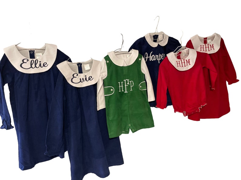 May include: Five children's clothing items on hangers. Two blue dresses with white collars, one green corduroy overall, one red long-sleeved shirt with a white collar, and one blue long-sleeved shirt with a white collar. The dresses and shirts are embroidered with names: Ellie, Evie, Harper, and HHM.