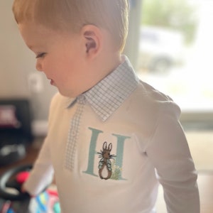 May include: A white long-sleeved shirt with a blue and white plaid collar. The shirt features an embroidered letter "H" with a small rabbit inside. The rabbit is brown with blue eyes and is standing on a green patch of grass.
