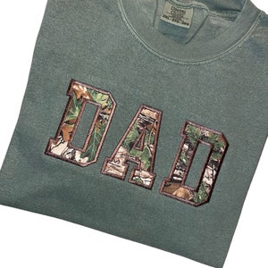 May include: A green t-shirt with the word "DAD" in large, white letters outlined in brown. The letters are filled with a camouflage pattern.