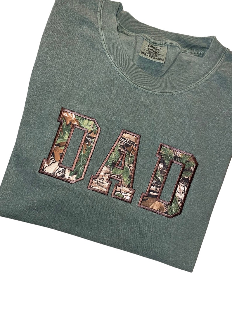 May include: A green t-shirt with the word "DAD" in large, white letters outlined in brown. The letters are filled with a camouflage pattern.