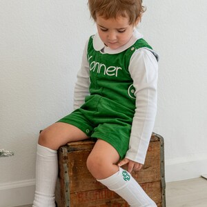 May include: A young child wearing a green corduroy overall with the name "Konner" embroidered on the front. The child is sitting on a wooden crate and wearing white knee-high socks with green embroidered initials.