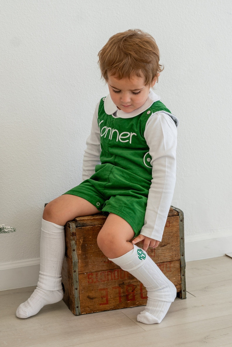 May include: A young child wearing a green corduroy overall with the name "Konner" embroidered on the front. The child is sitting on a wooden crate and wearing white knee-high socks with green embroidered initials.