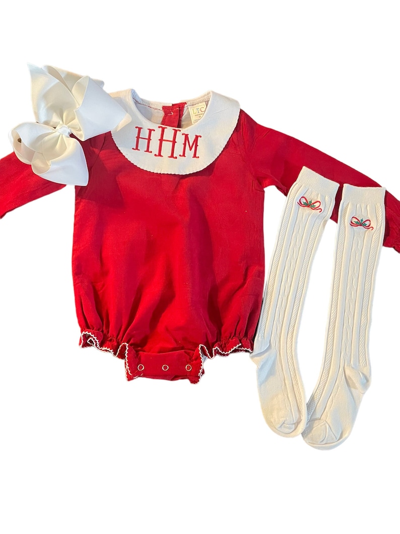 May include: Red long-sleeved baby onesie with a white Peter Pan collar and the monogram "HHM" embroidered on the front. The onesie has a white ruffle trim and is paired with white cable knit socks with a small embroidered bow and holly design.