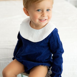 May include: A young child wearing a navy blue dress with a white collar and white ruffled sleeves. The dress has a white trim around the bottom. The child is wearing a white bow headband and has blonde hair. The child is sitting on a light blue stool.